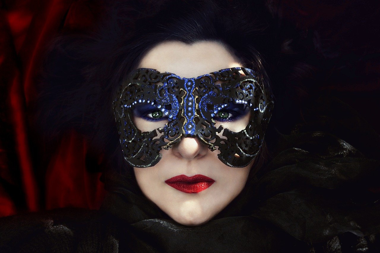 Pale woman with red lipstick and dark eyeliner wearing a blue and black masquerade mask. Red satin in background