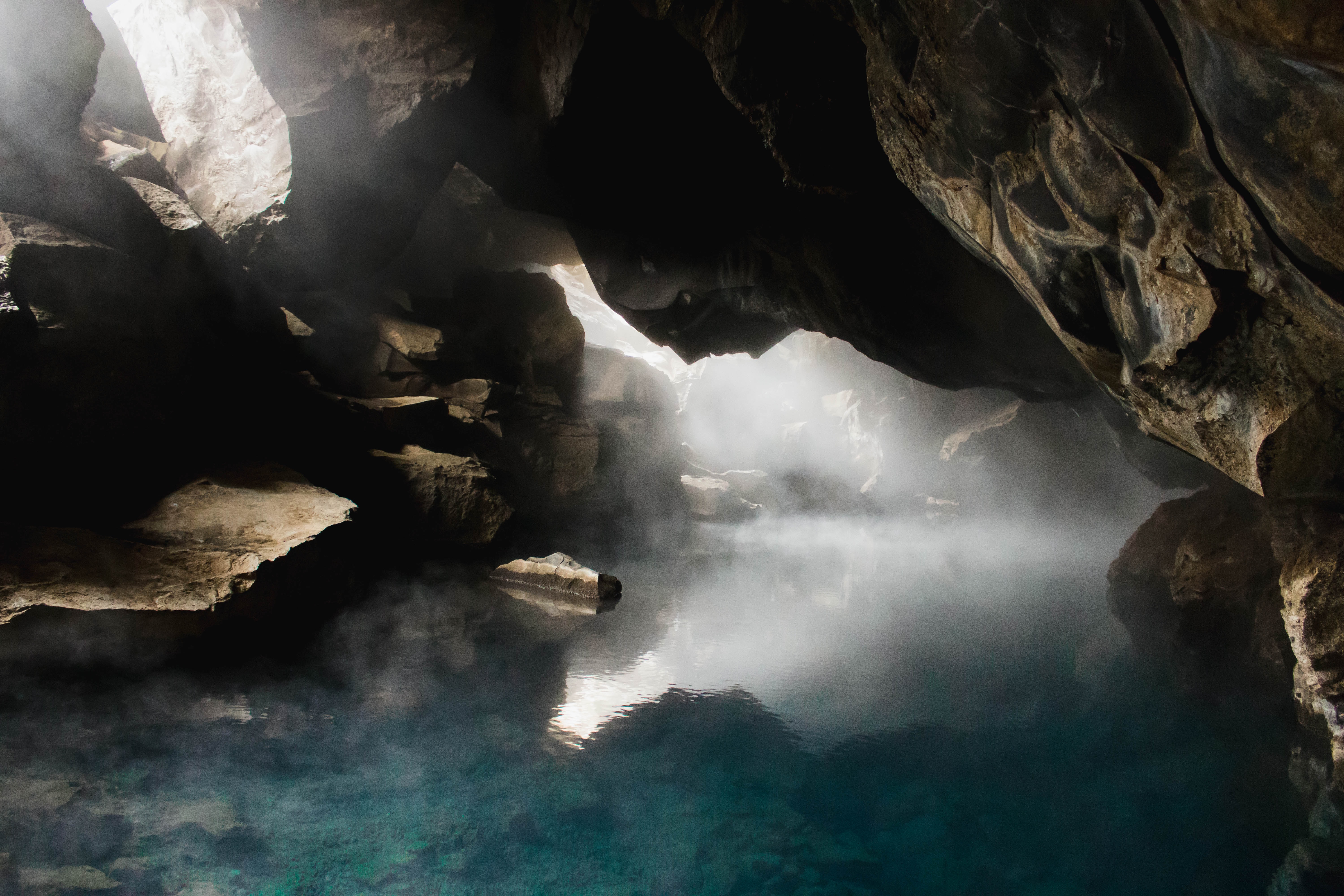 Photography of a cave with light shining from holes up above, illuminating the mist below. 