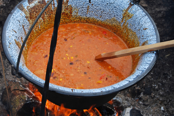 Top down view of a tomato based stew with beans and vegetables hung on an iron tripod, cooking over a campfire.