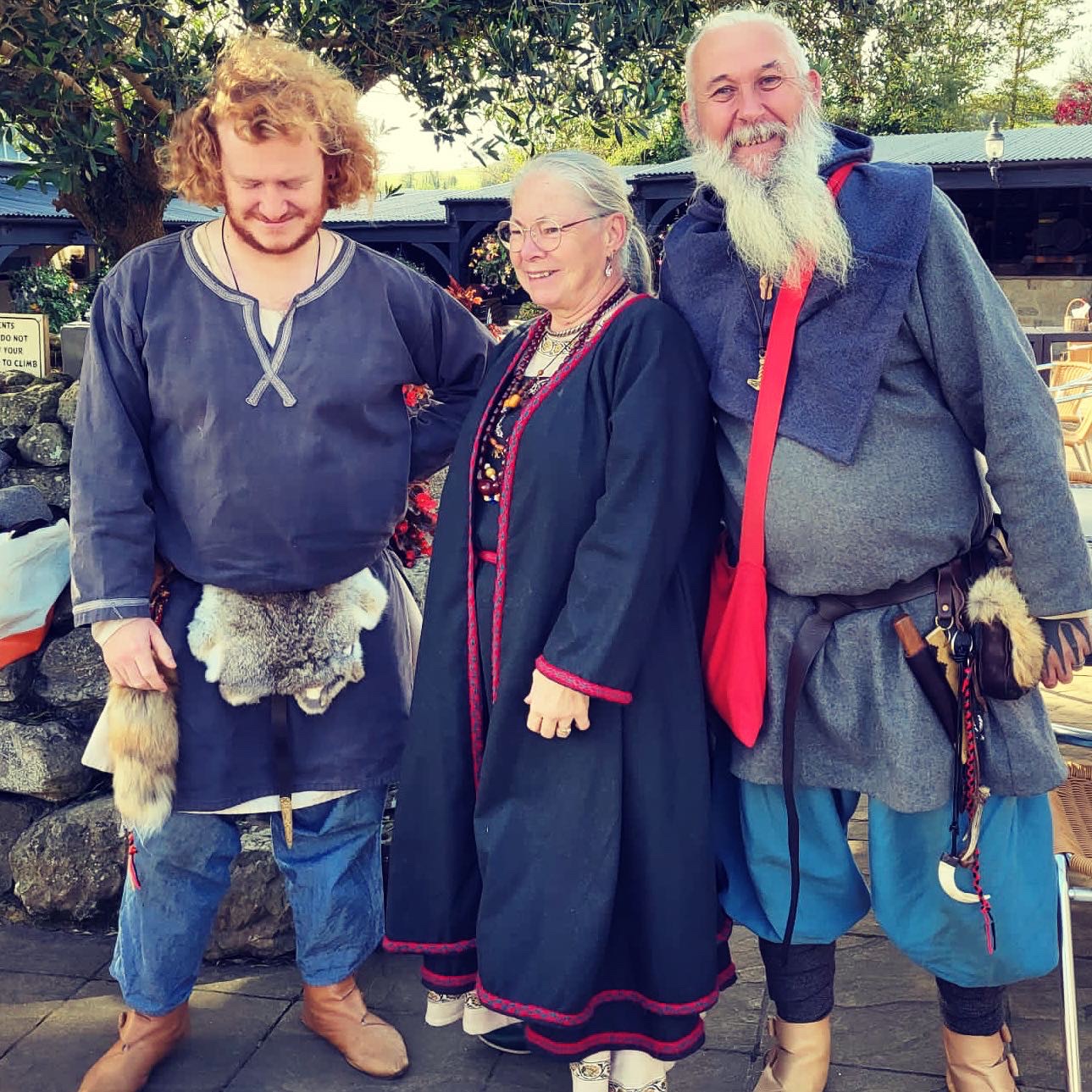 Three Viking reenactment players, Rekr, Mutter Maia, and Habard stand together by a tree. They wear traditional garb and accessories in blues and greys, with flashes of bright red. Hanging from their belts can be seen weapons, pouches, tools, and furs.