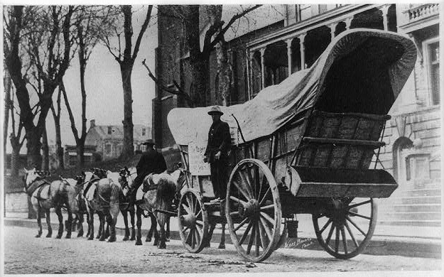 Photograph shows a team of six horses with covered wagon on street; man sits on back of horse and another man standing on sideboard.