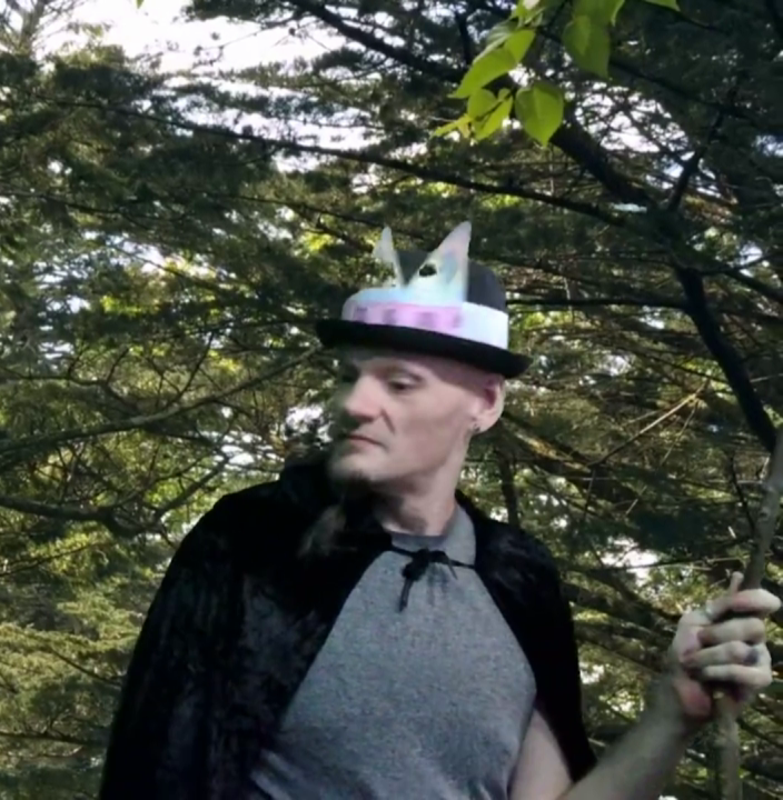 A man in a paper crown and robe, staff in hand, with a background of willow trees