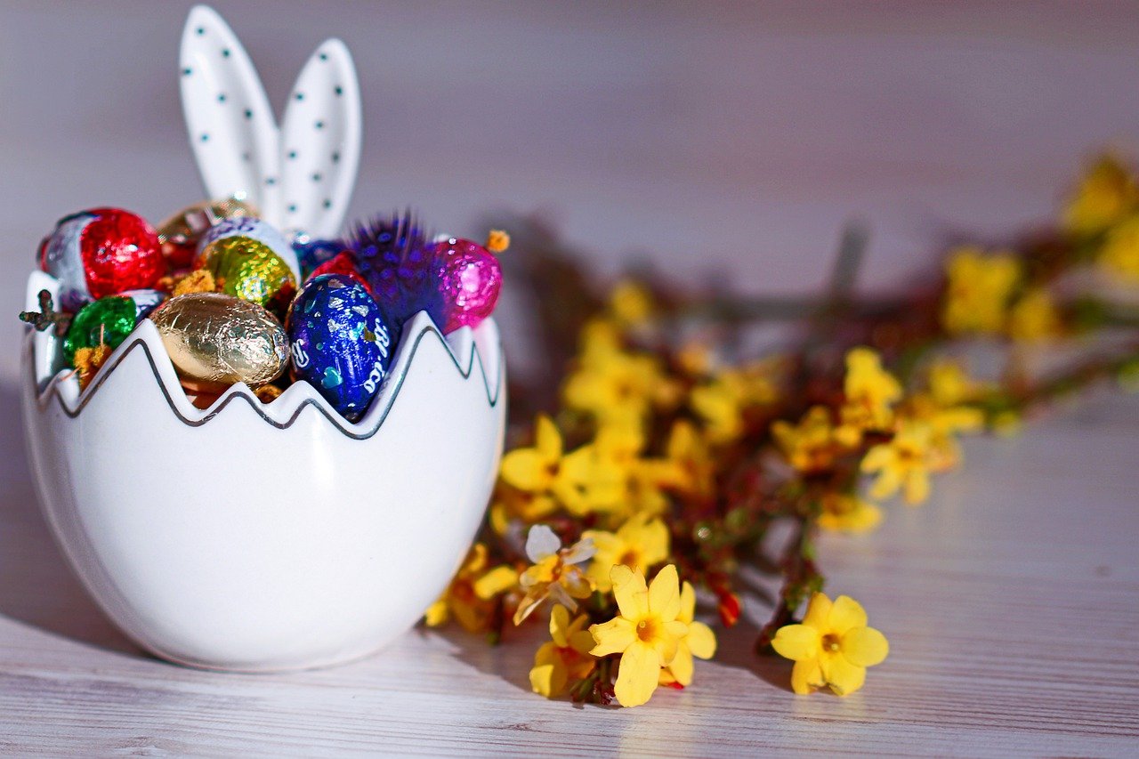 A ceramic half-egg and bunny ears, filled with bright-wrappered chocolate eggs, next to yellow flowers