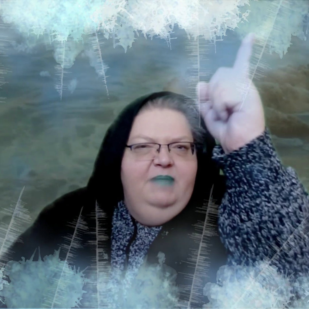 A woman in a black shawl and teal lipstick, with frost in the foreground and ocean in the background, lifting her finger to the air with a stern expression on her face