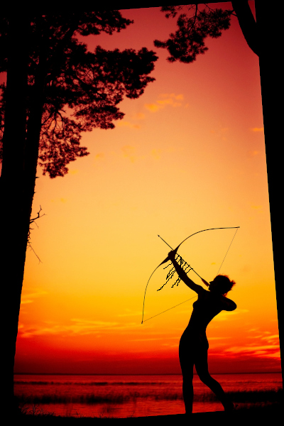 Beautiful huntress at sunset stock photo framed by two tall trees. The orange sun is setting over a shallow body of water with grasses and reeds sticking out of it.